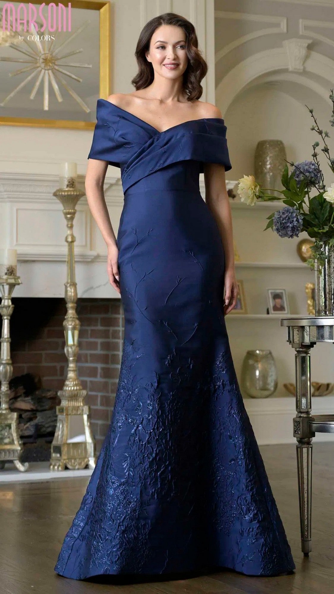 A woman in the navy Marsoni MV1378 long formal dress with an off-shoulder neckline and floral texture stands in a decorated living room.